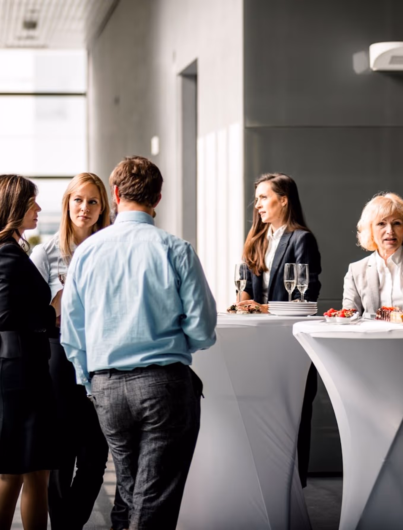 Group of professionally dressed people networking at a cocktail event with white high tables and drinks.