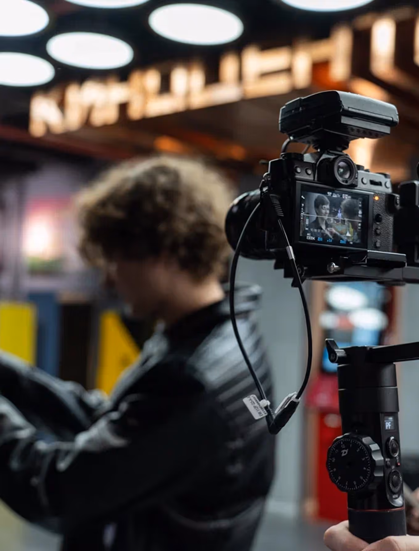 Close-up of camera on gimbal filming a person with curly hair wearing a dark jacket in a blurred background.
