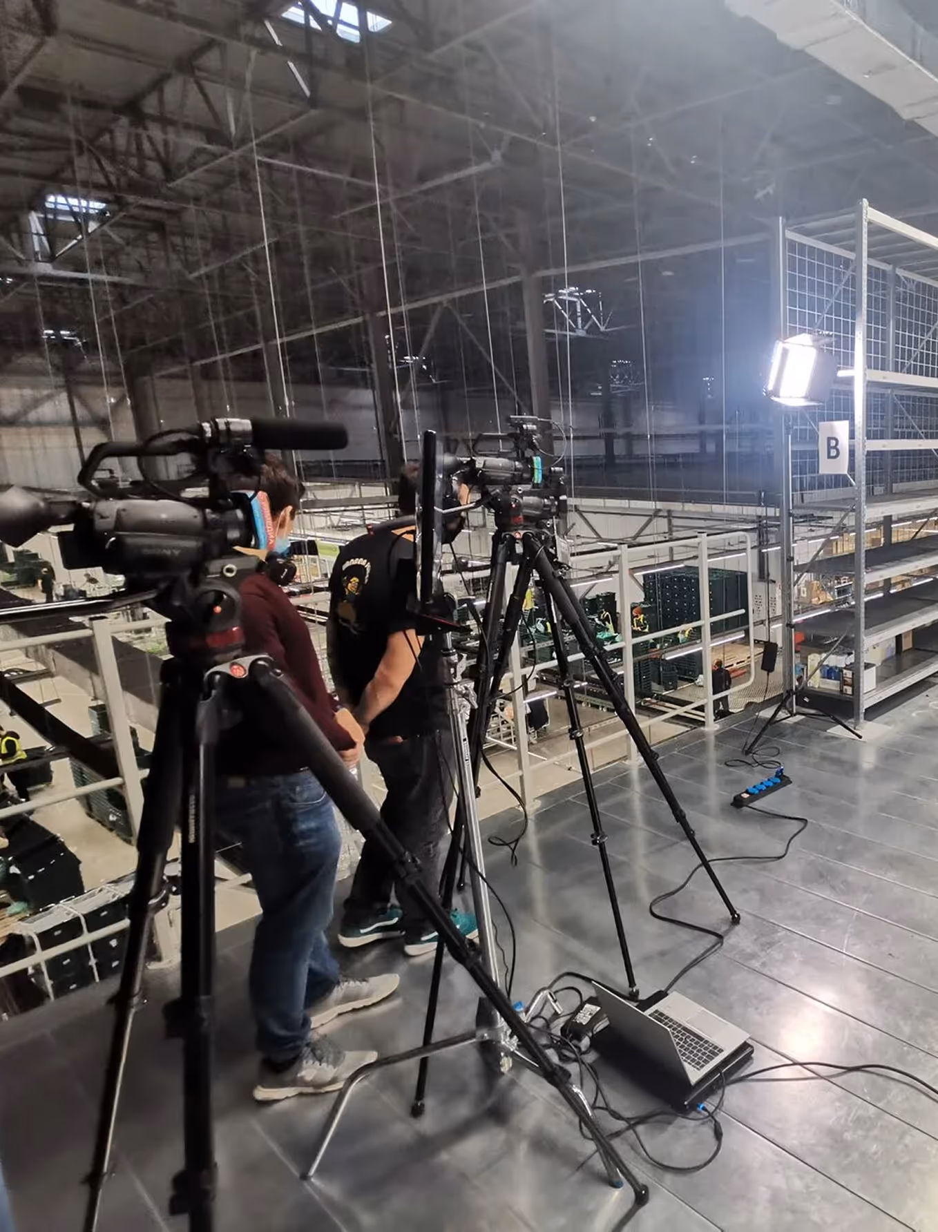 Two videographers operating cameras on tripods with lighting setup in an industrial warehouse.