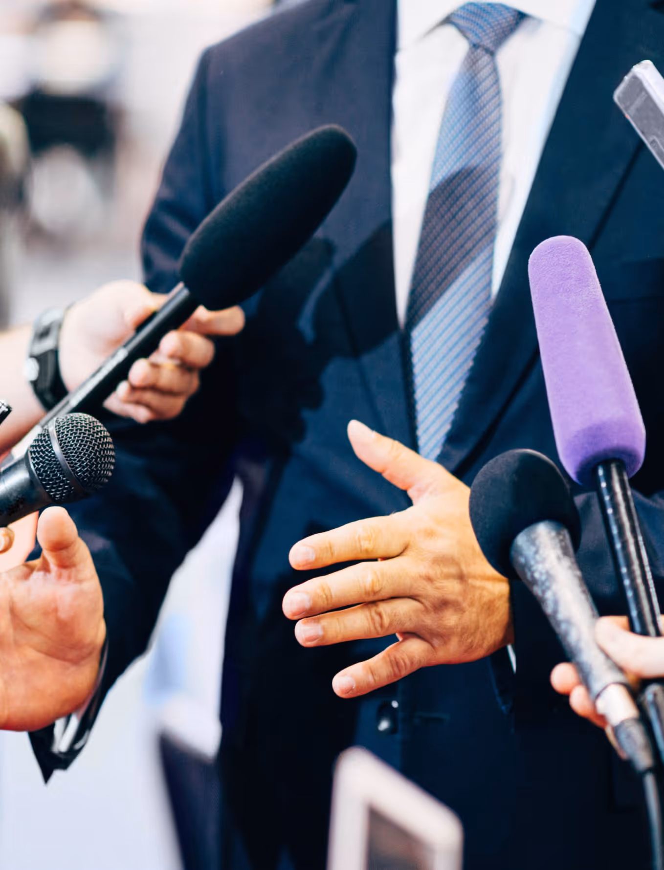 Person in suit and tie speaking to multiple microphones held by reporters.