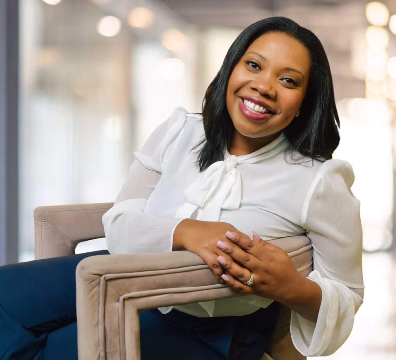 Smiling woman in white blouse sitting in beige chair with bright blurred background.