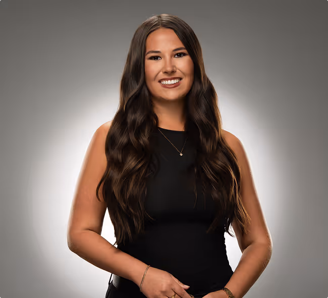 Young woman with long wavy hair wearing a black top and a pearl necklace looking at the camera.