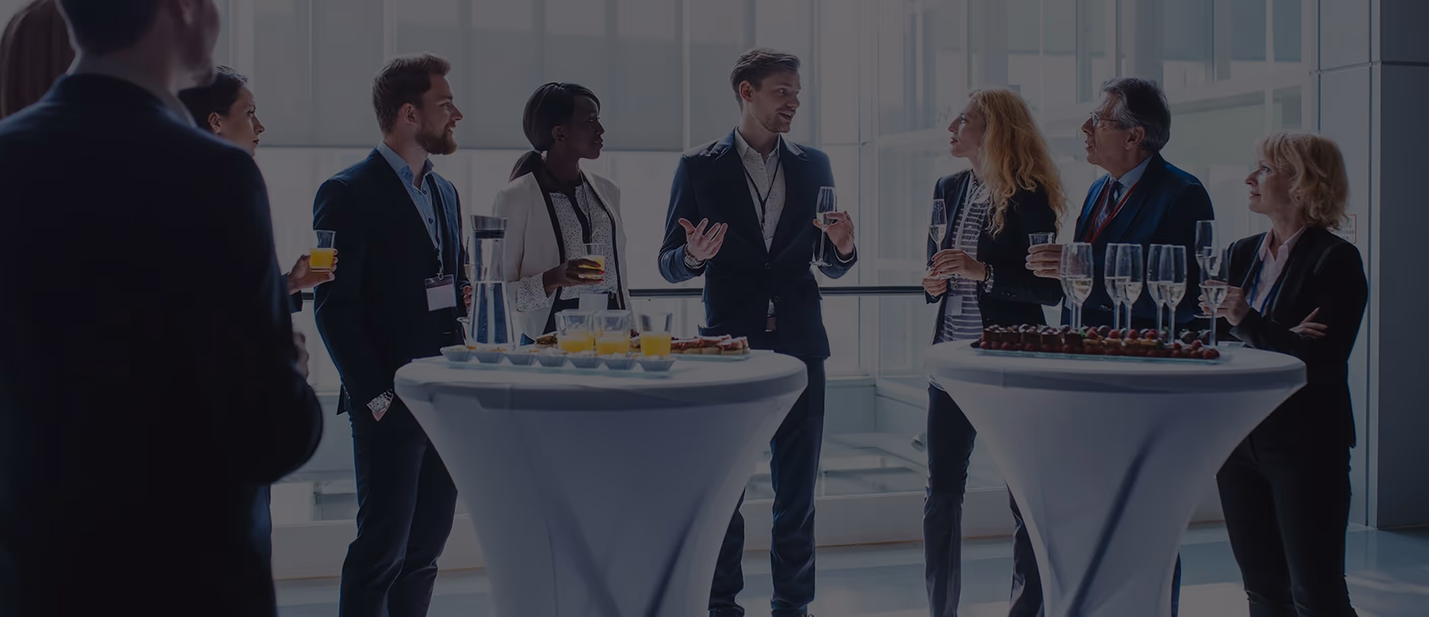 Group of business professionals chatting around cocktail tables with drinks and appetizers at a networking event.