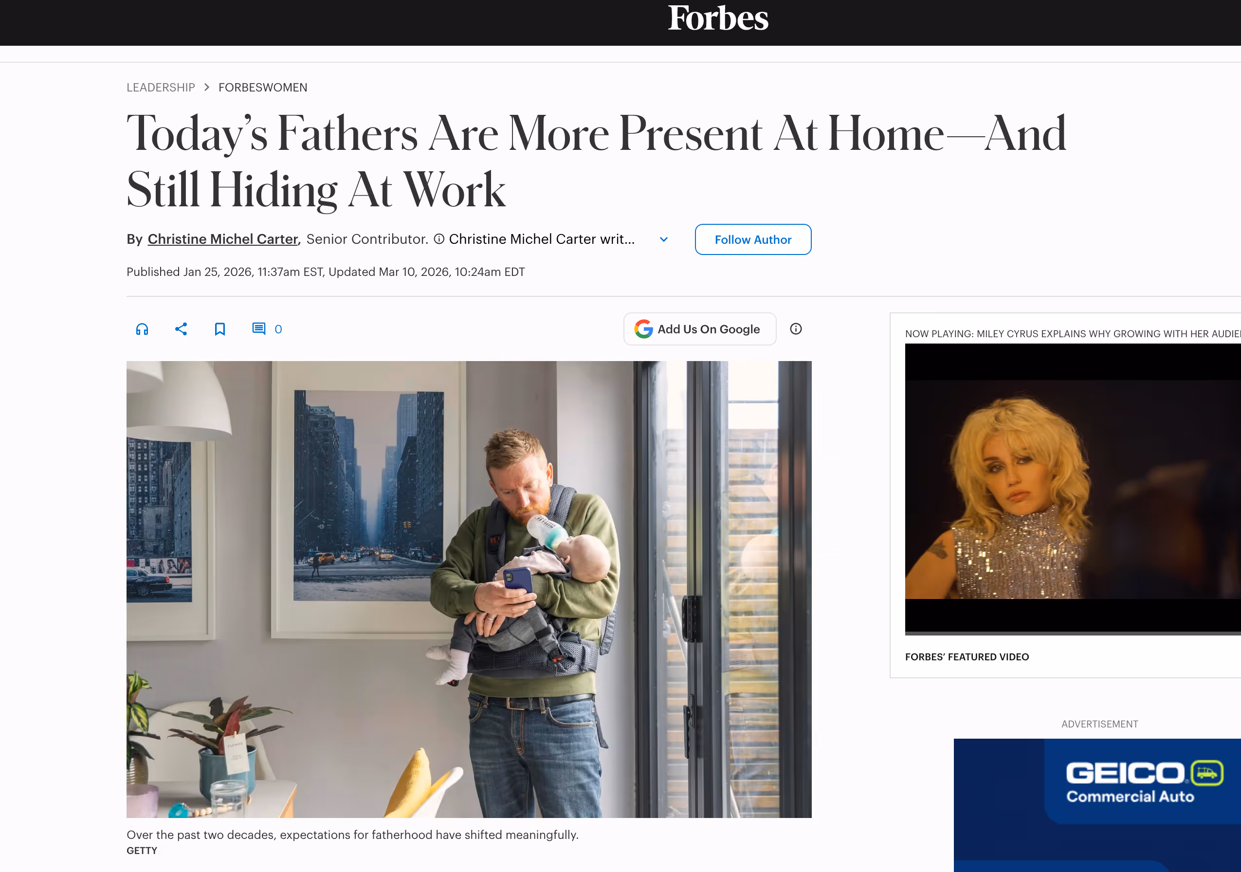 Father feeding baby with bottle while holding a phone at home near a window and city photos.
