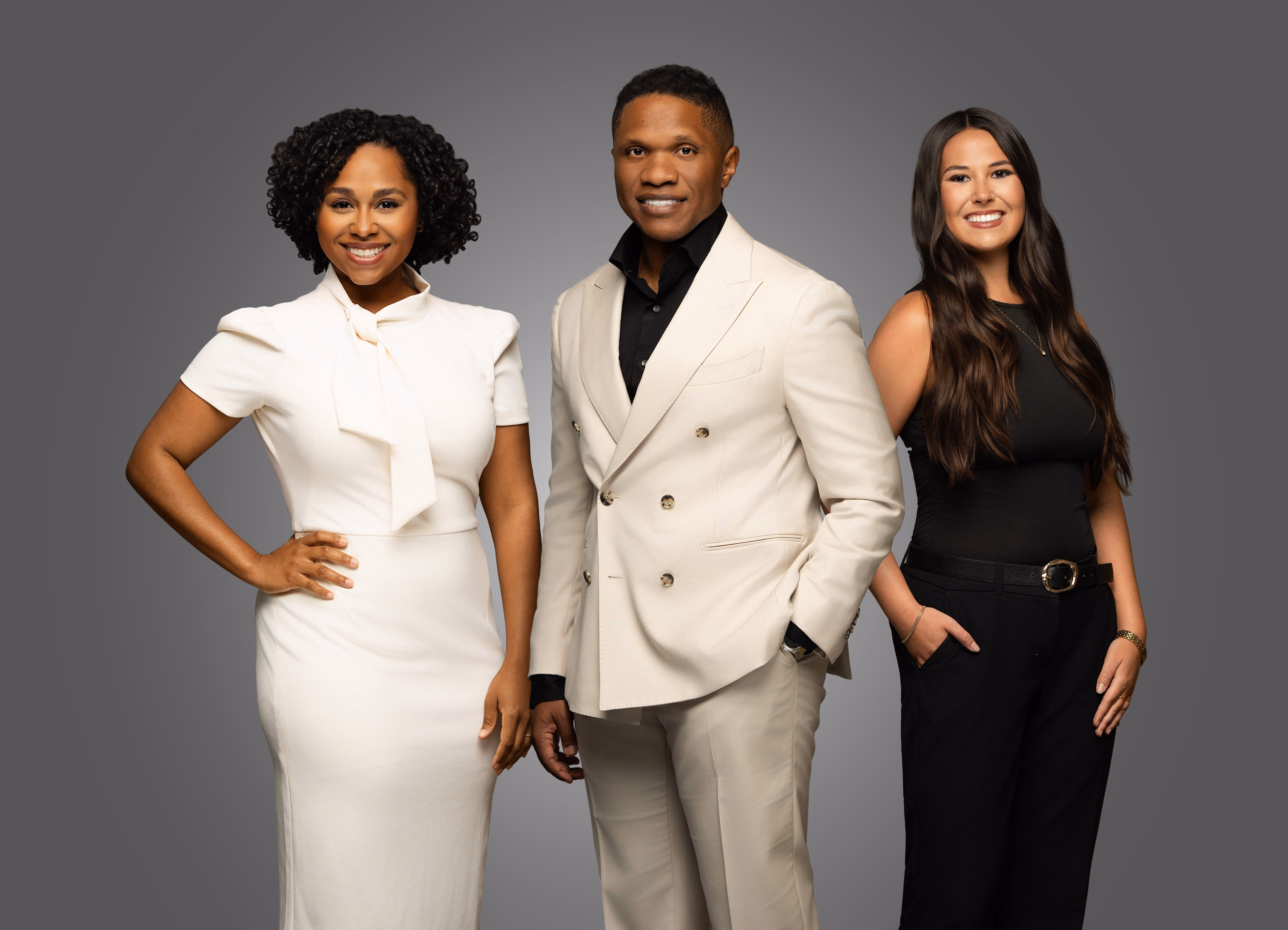 Four diverse professionals smiling, standing against a plain gray background in business attire.