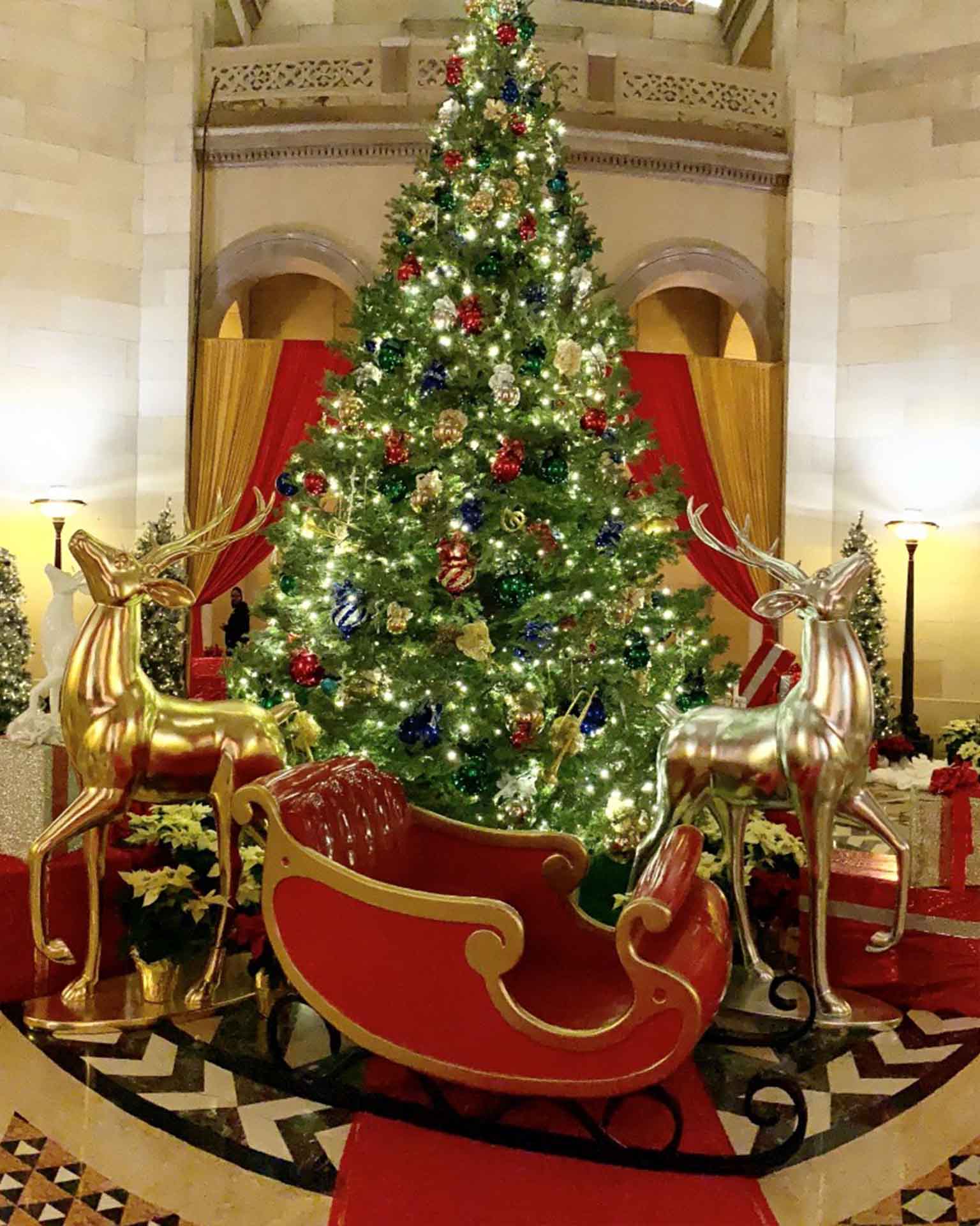 Decorated Christmas tree with lit ornaments beside red sleigh and gold and silver reindeer statues.