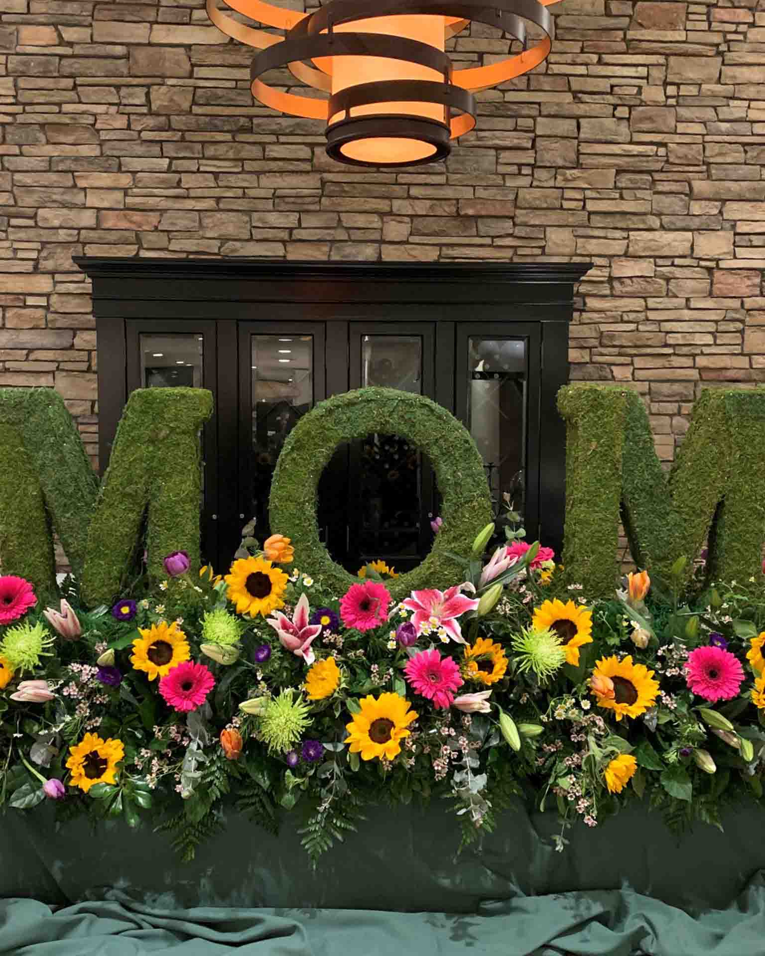 Green moss letters spelling MOM adorned with sunflowers, pink lilies, and other colorful flowers.