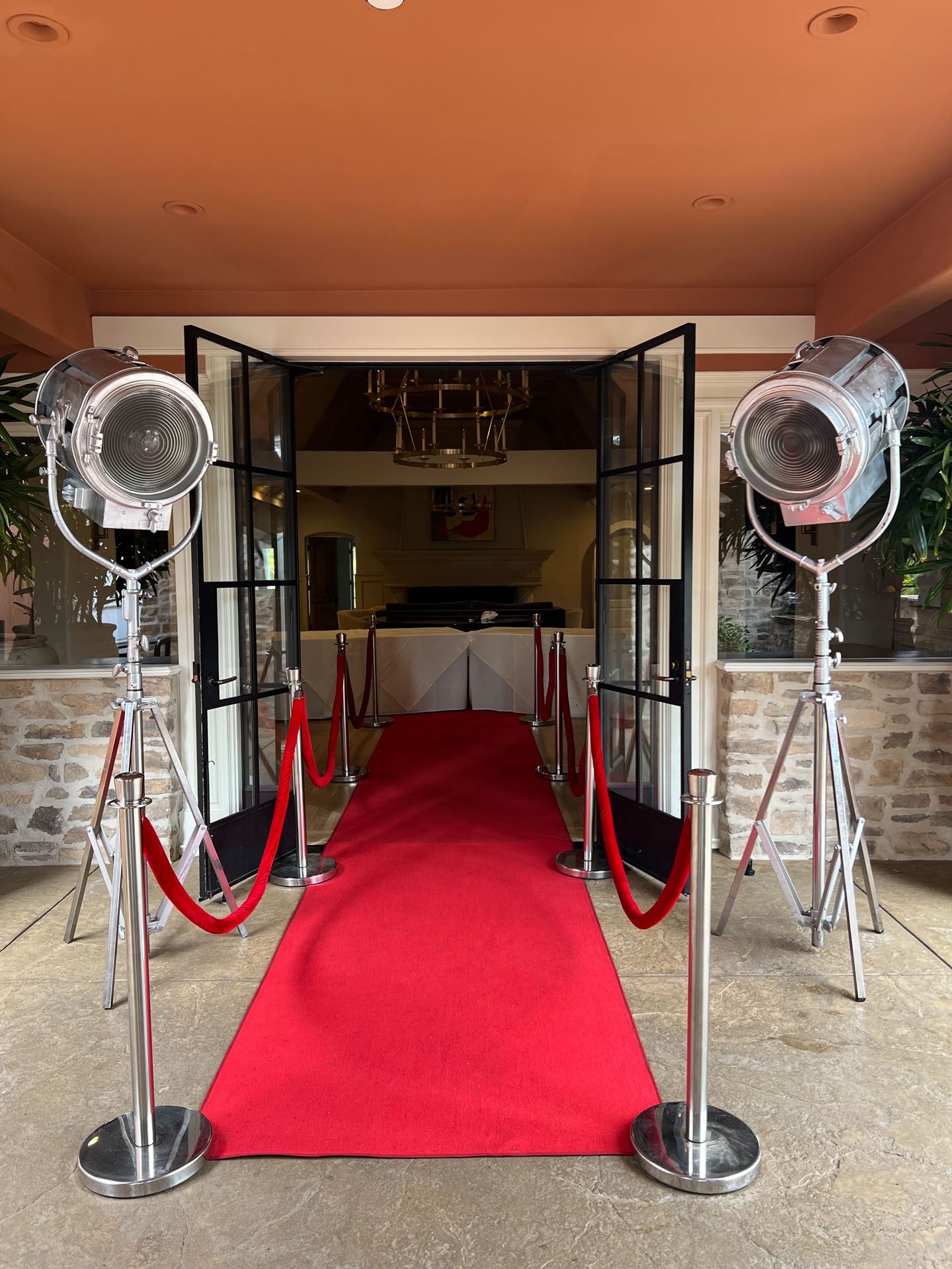 Hollywood style red carpet flanked by velvet ropes and large vintage studio lights at entrance of a room with stone walls.