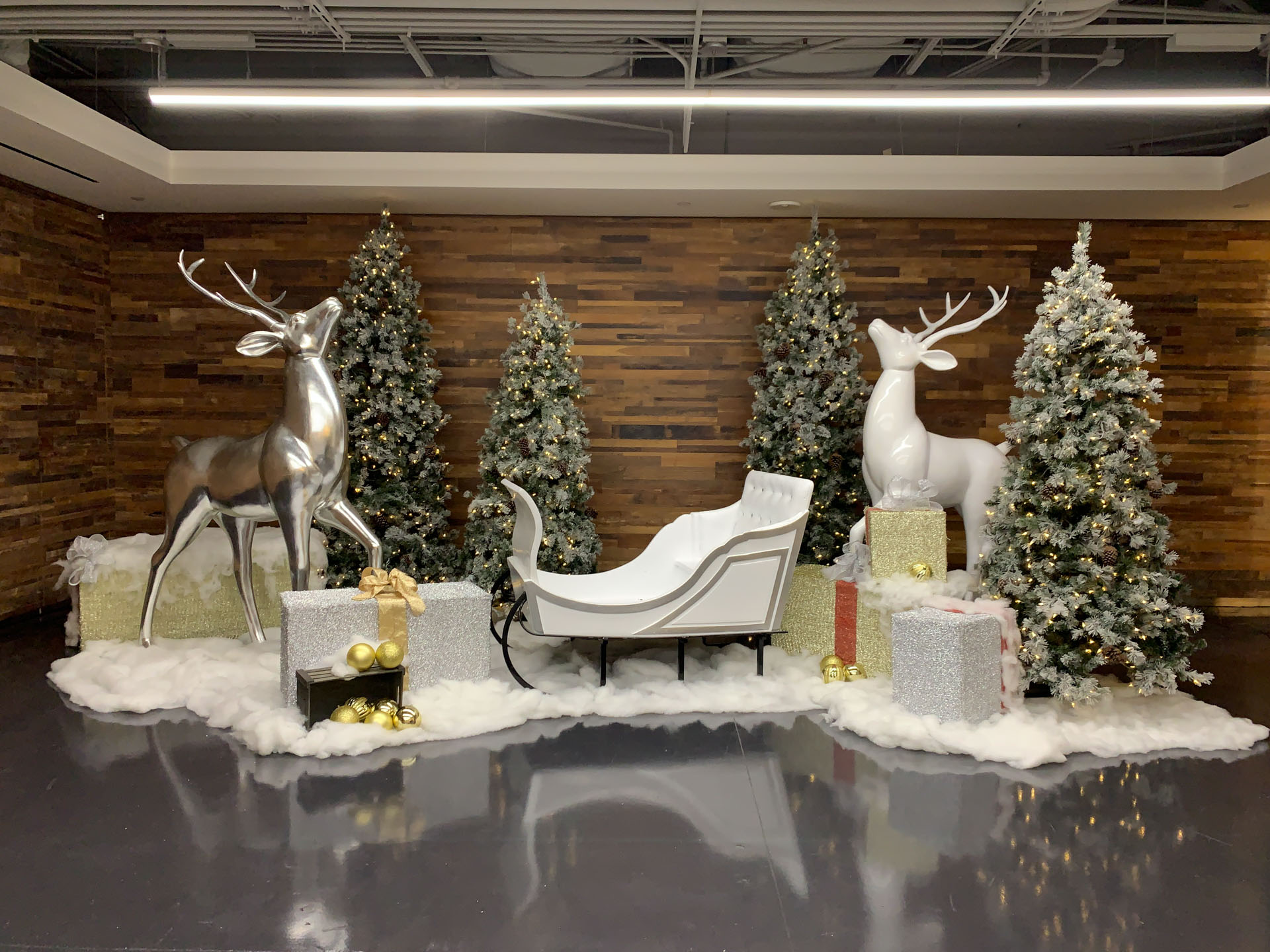 Holiday scene with white sleigh, silver and white reindeer statues, decorated pine trees, and gift boxes.