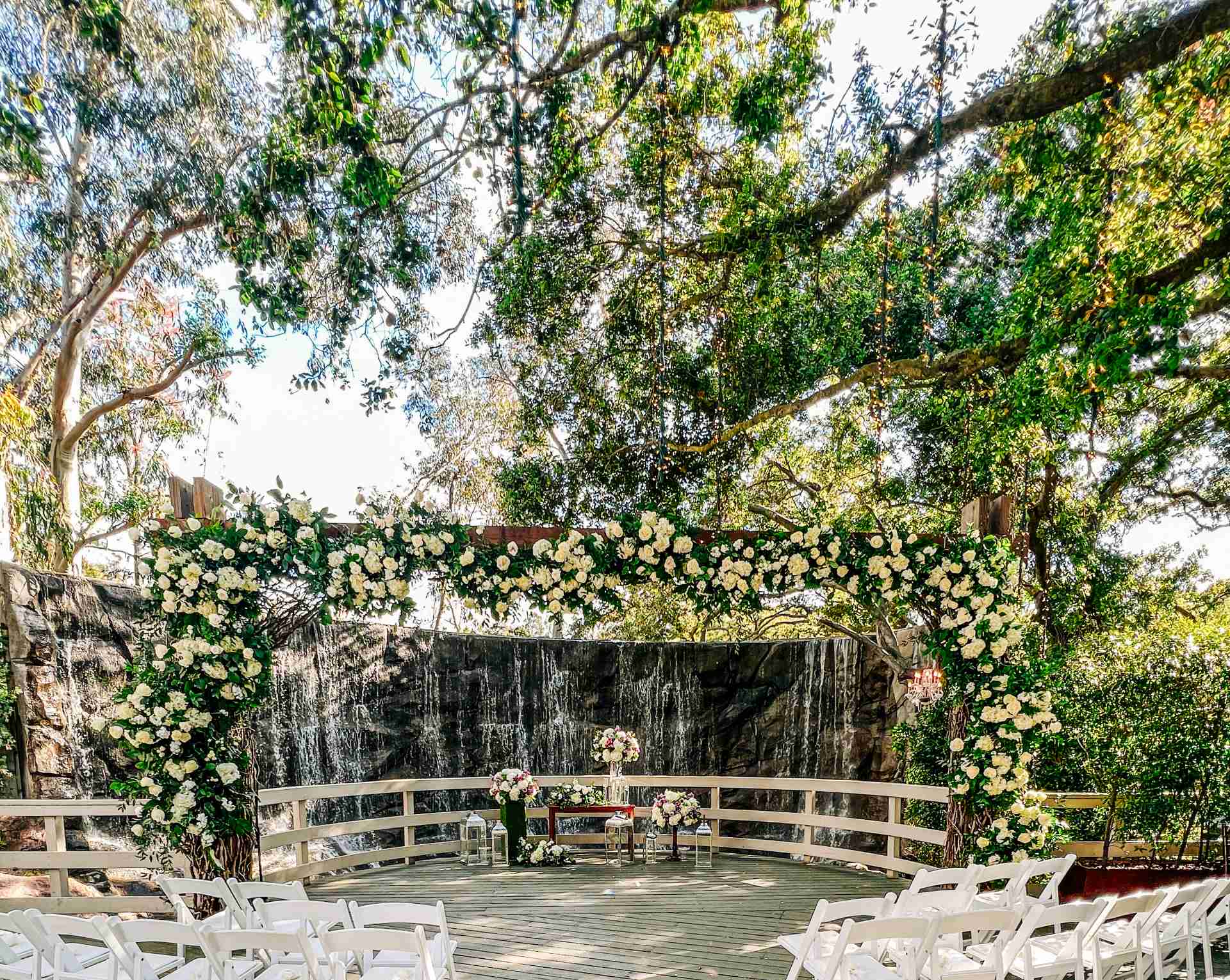 Outdoor wedding setup with white chairs, floral arch, and waterfall backdrop surrounded by trees.