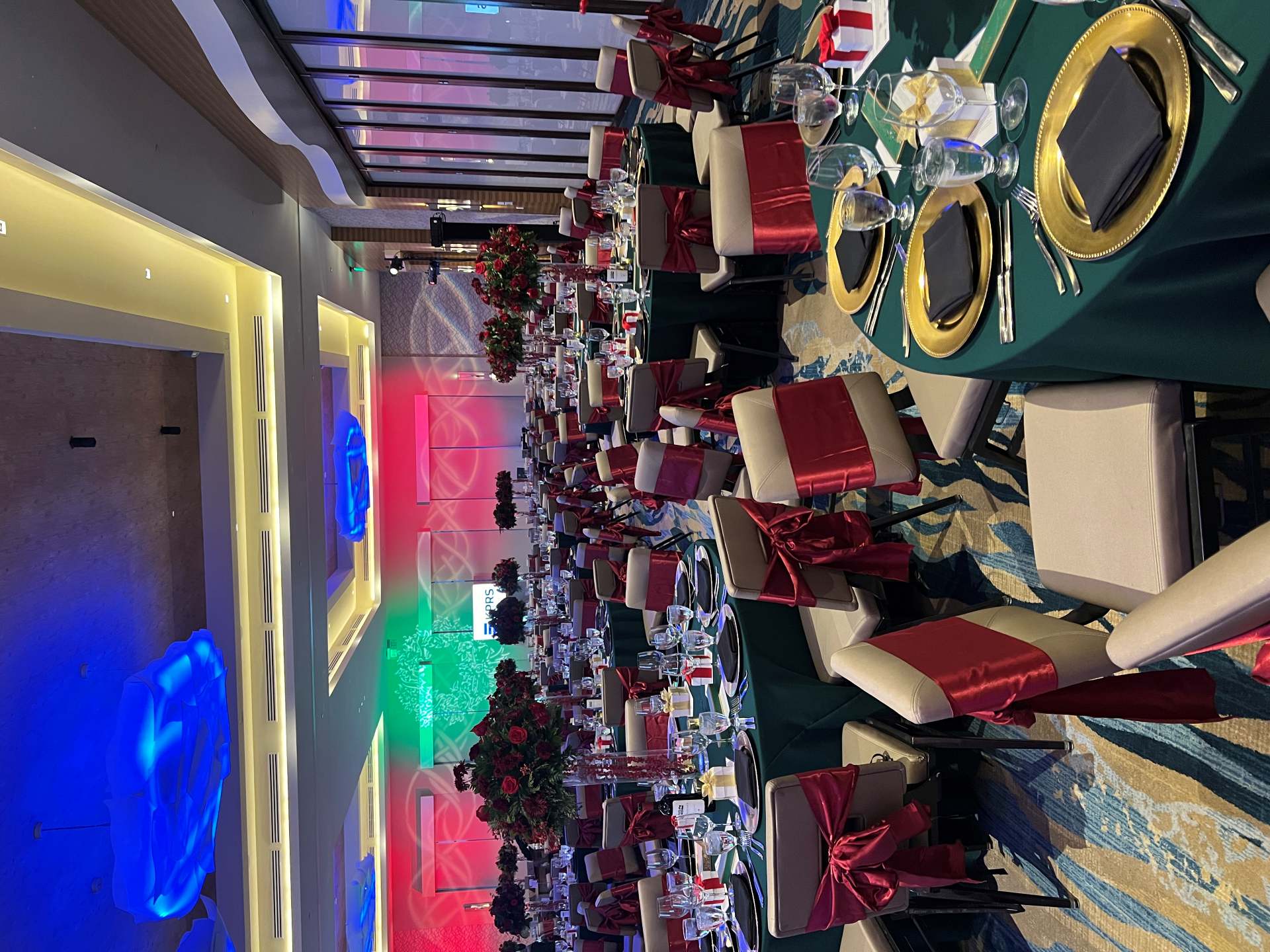 Large conference venue hall filled with dinner tables decorated with Christmas colors with green table cloths and red bows tied around each chair. 
