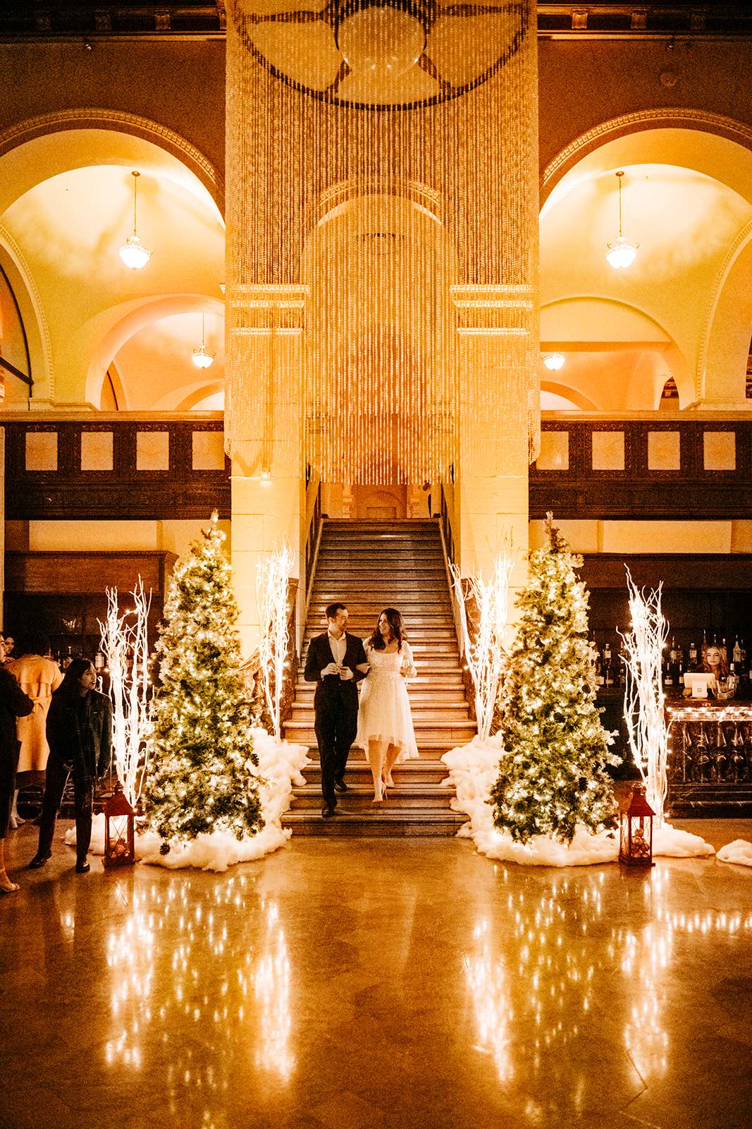Couple walking down a grand staircase between illuminated Christmas trees and glowing white branches inside an elegant venue.