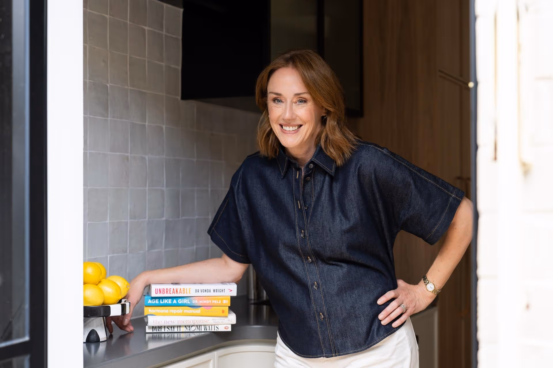 Smiling woman in a dark button-up shirt leaning on a kitchen counter with a stack of health-related books and a bowl of lemons.