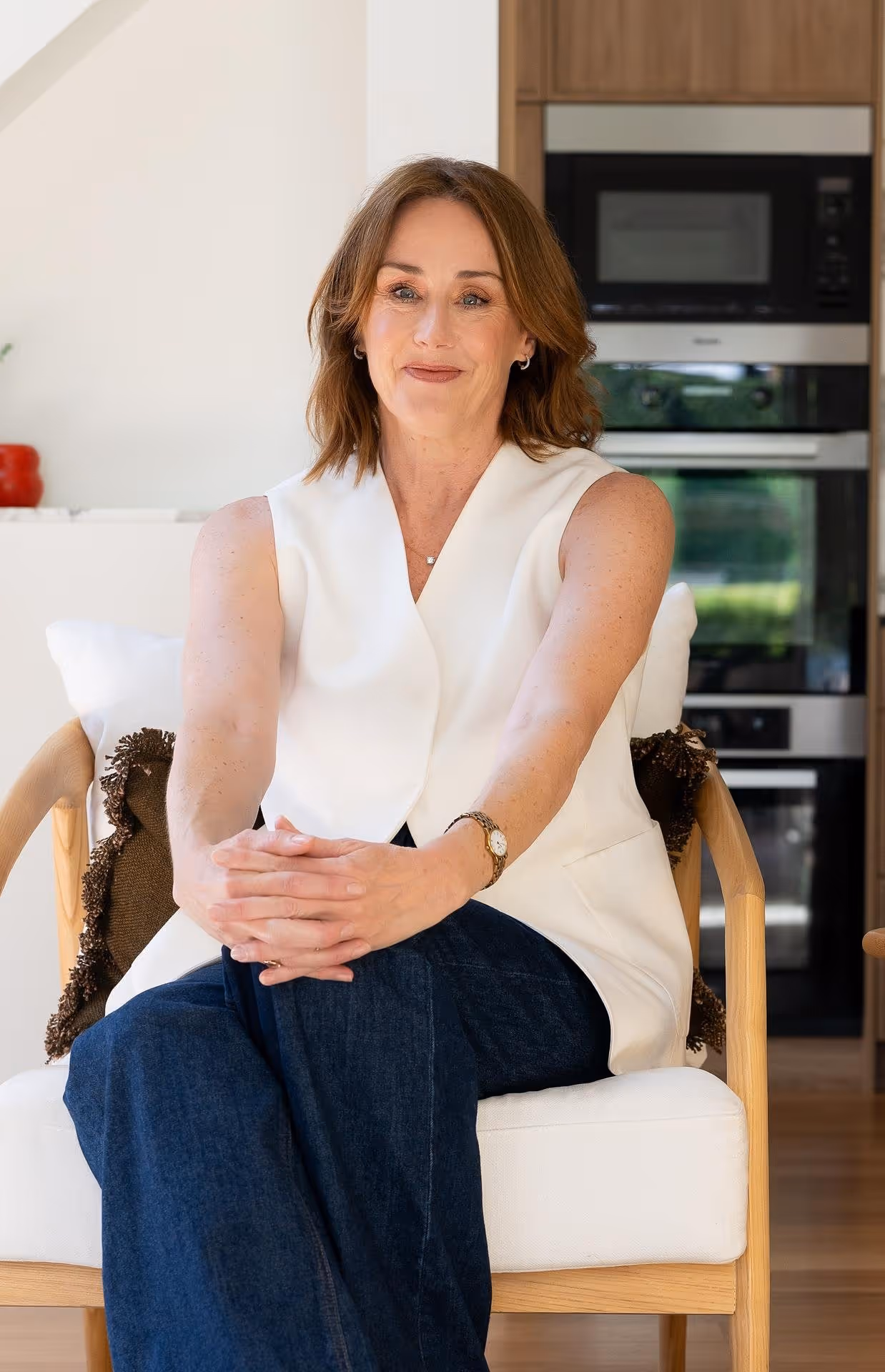Woman with shoulder-length brown hair sitting with legs crossed on a wooden chair wearing a sleeveless white top and dark jeans in a modern kitchen.