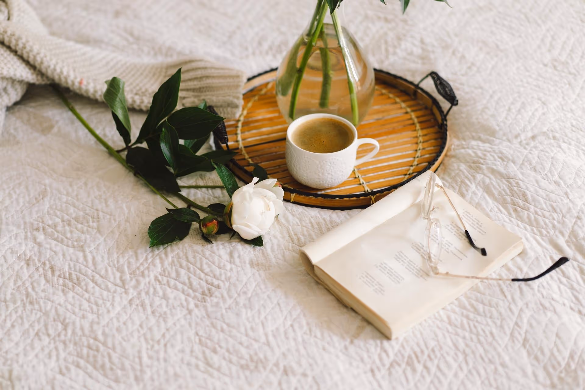 White coffee cup on a bamboo tray next to a white rose and an open book with reading glasses on a quilted white bedspread.