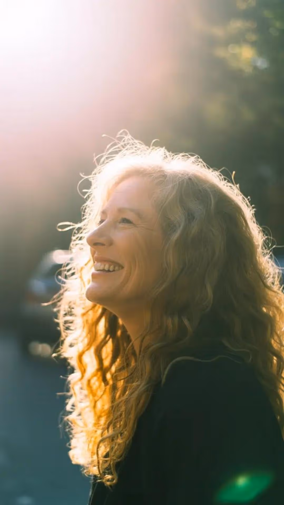 Smiling woman with curly blonde hair illuminated by sunlight, looking up outdoors.