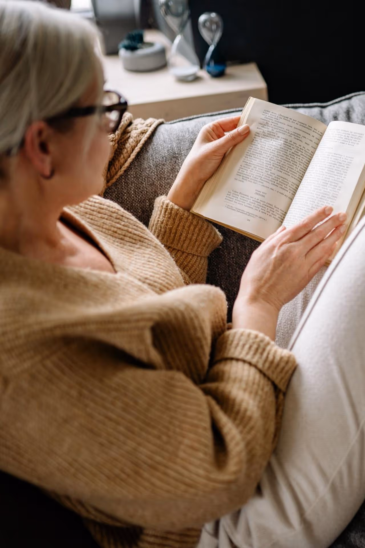 Person in a beige sweater and glasses sitting on a gray couch reading a book.