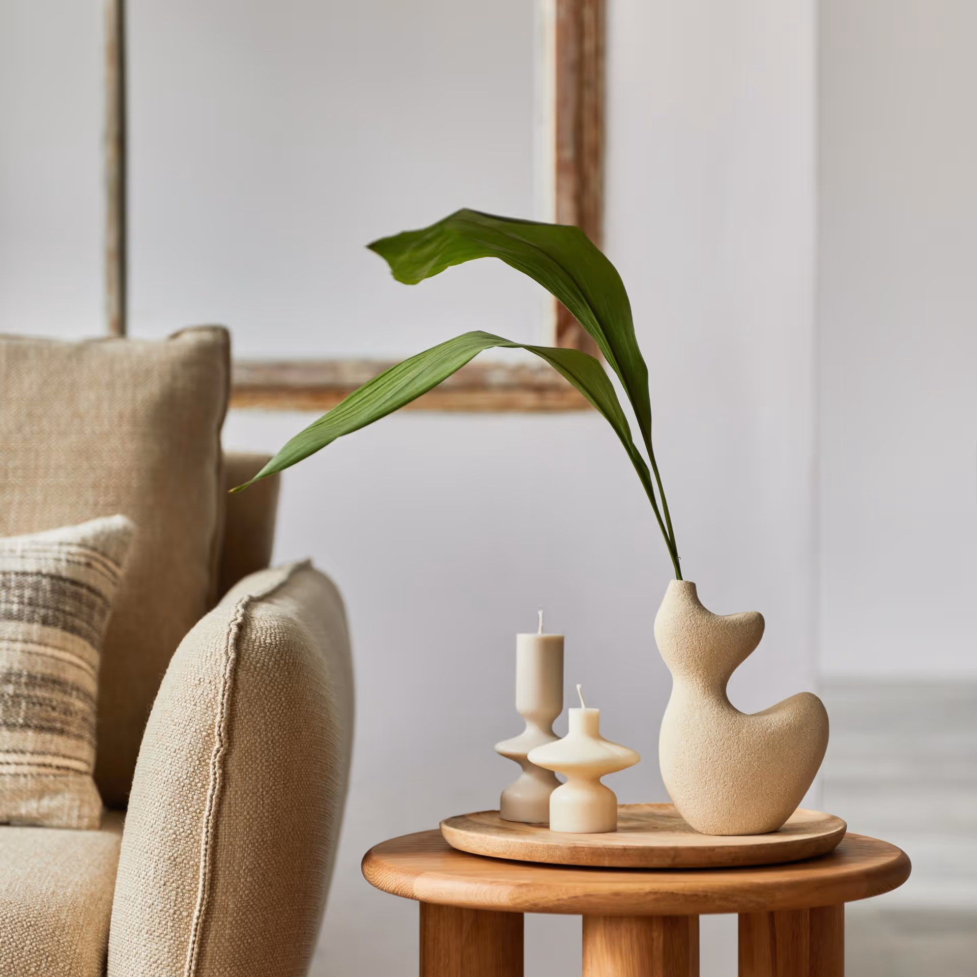 A minimalist living room features a sculptural beige vase with a green leaf on a round wooden table. Two uniquely shaped candles add a calming touch.