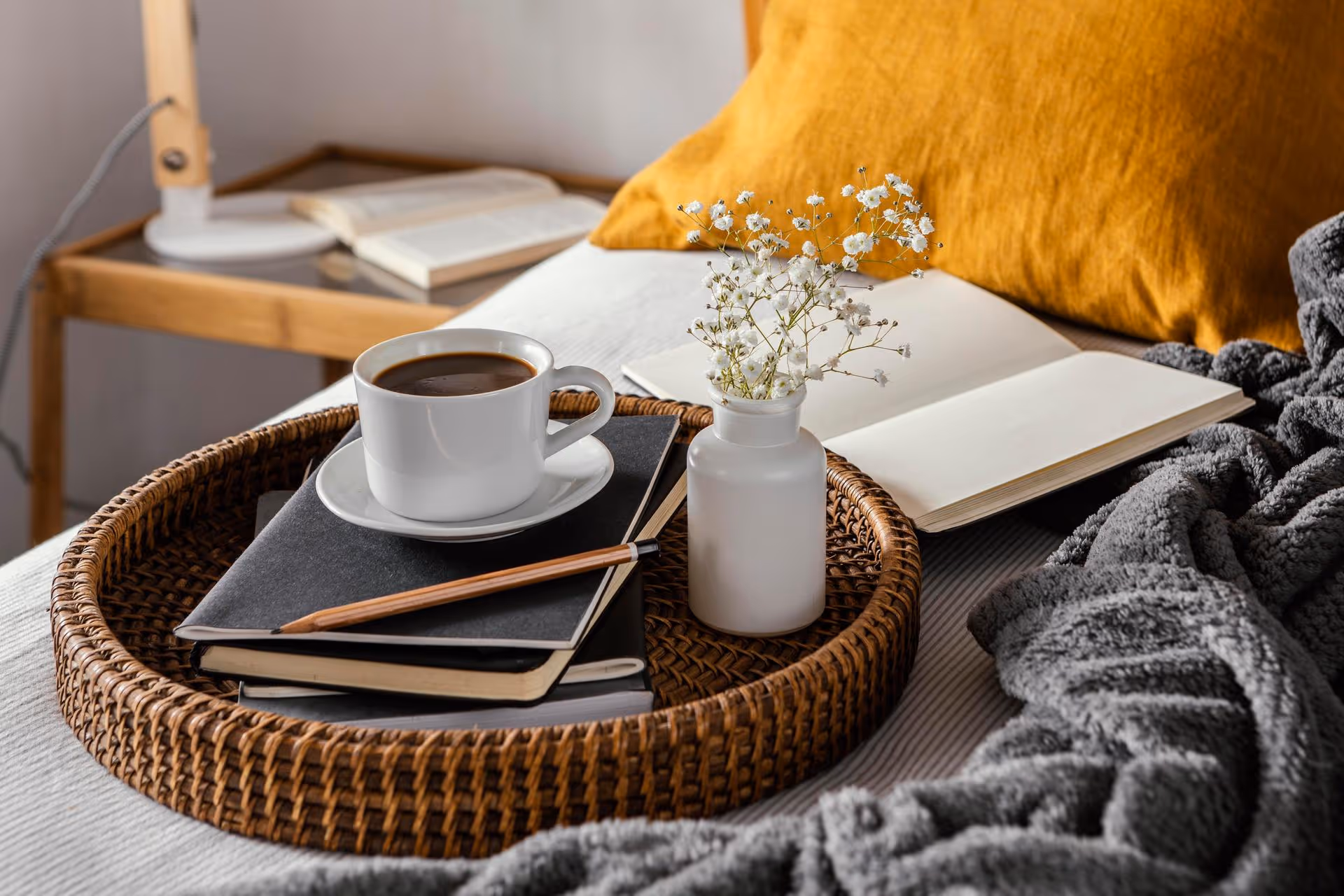 A wicker tray on a bed holds a cup of coffee, a pencil, two notebooks, and a small vase with white flowers; an open book and lamp are in the background.