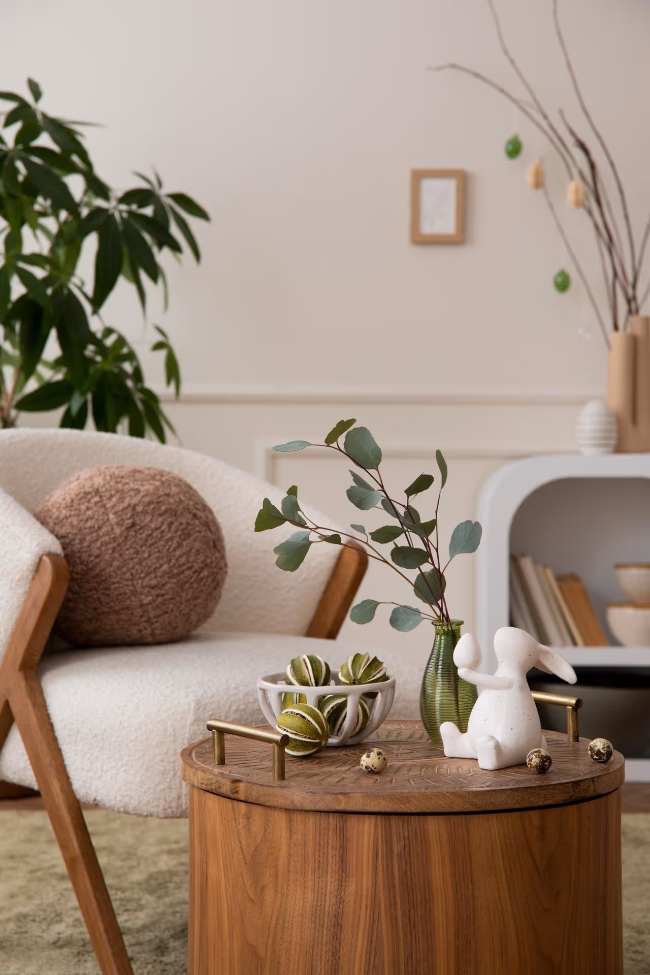 Cozy living room with a white chair and brown cushion beside a wooden table. Table holds a vase, ceramic bunny, and decorative bowl. A plant is in the background.