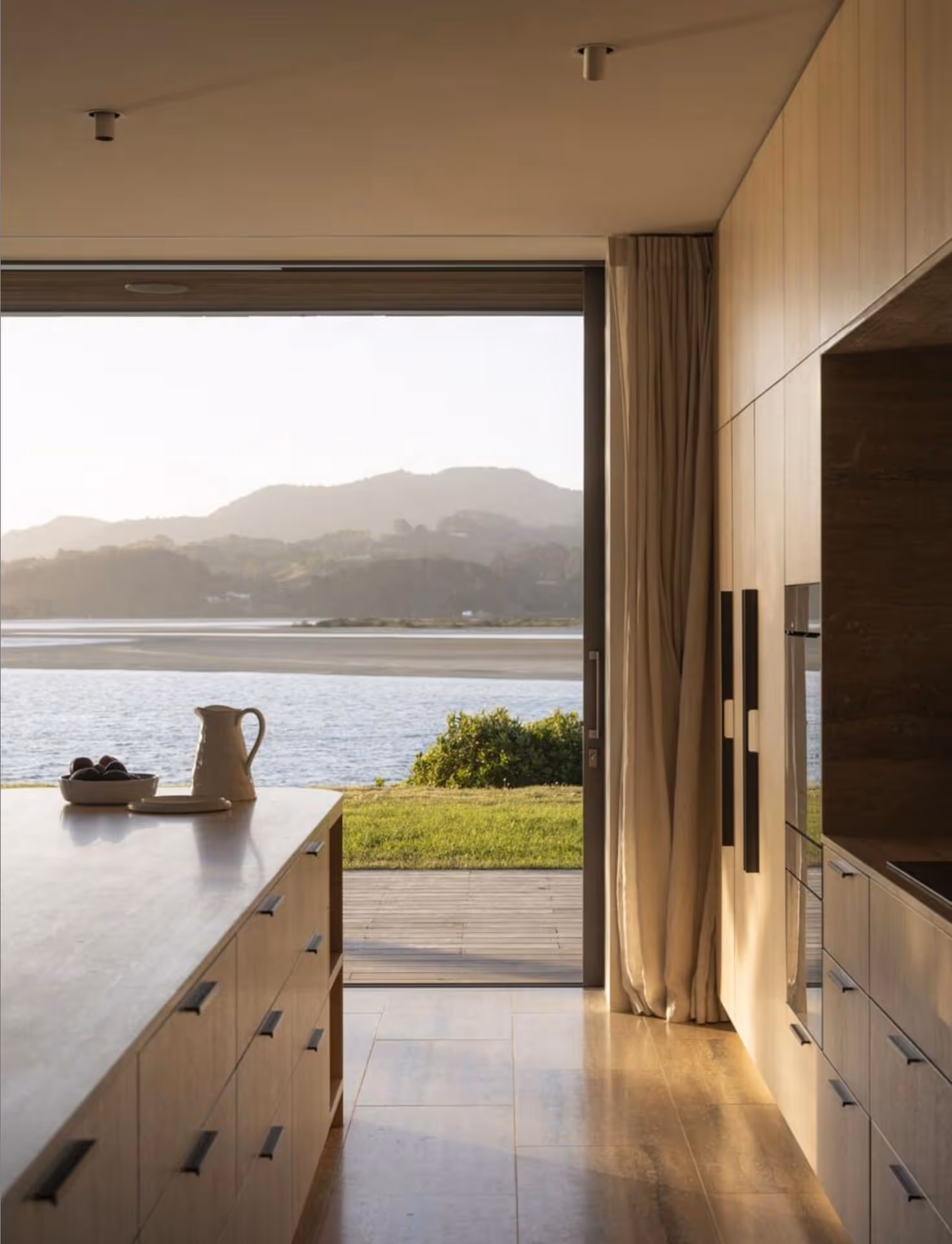 Modern kitchen with wooden cabinets, large island featuring a white pitcher, and floor-to-ceiling window revealing a serene lake and mountain view.