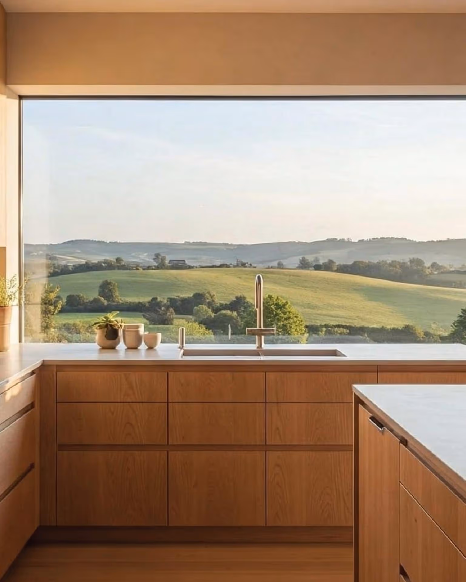 Modern kitchen with wooden cabinets and a large window showcasing a serene countryside landscape. Light floods in, creating a warm and inviting atmosphere.