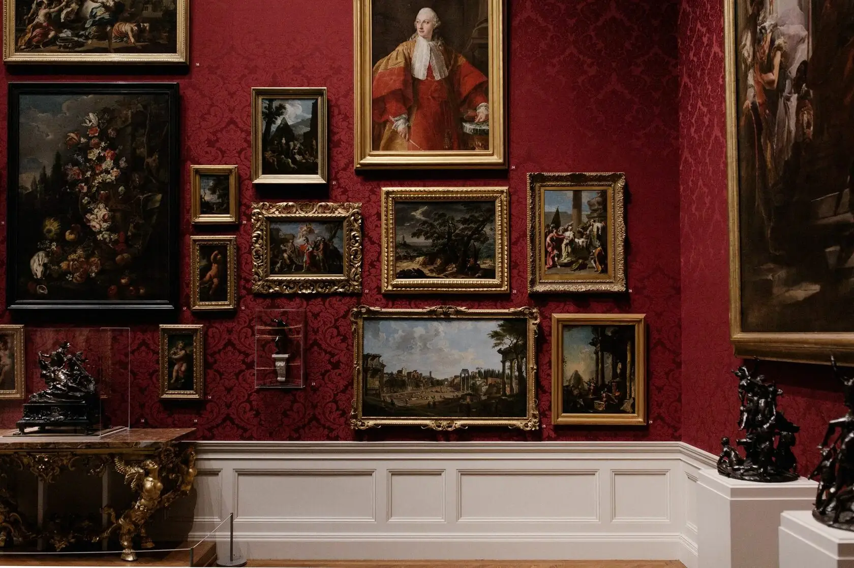 Victorins - gallery wall with classical paintings in ornate frames on red wallpaper above white wainscoting