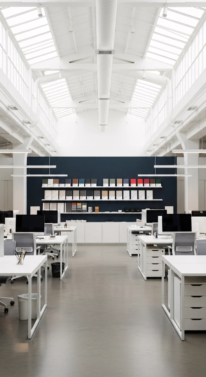 Bright modern office with rows of white desks, computers, gray chairs, and a book display on a dark wall.
