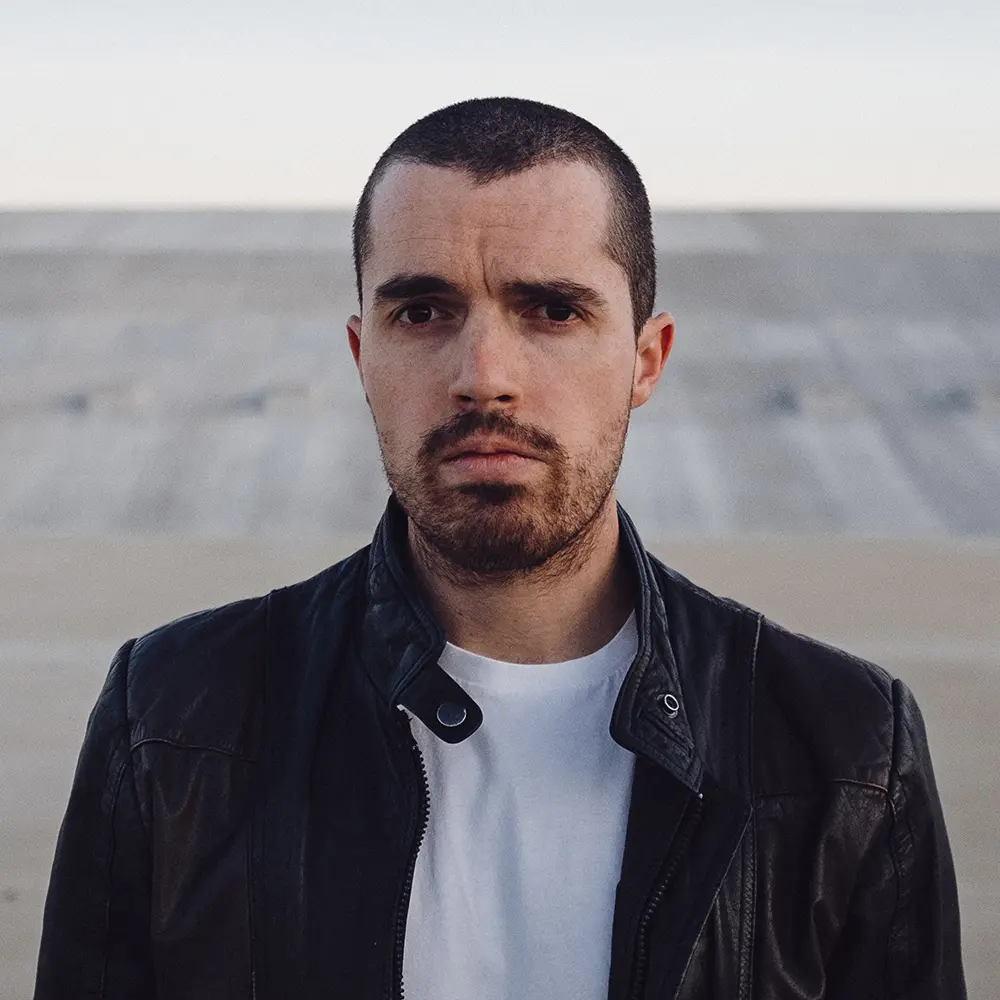 Serious man with short hair and beard wearing a black leather jacket and white shirt outdoors.