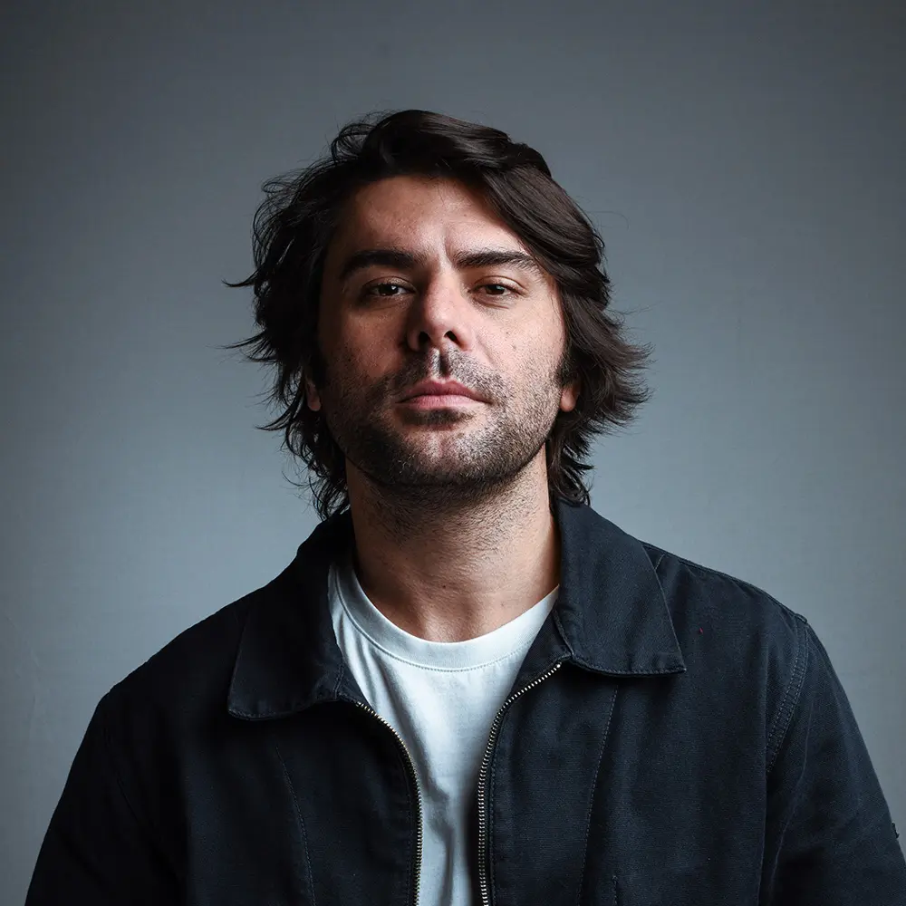 Man with medium-length dark hair and beard wearing black jacket and white shirt against gray background.