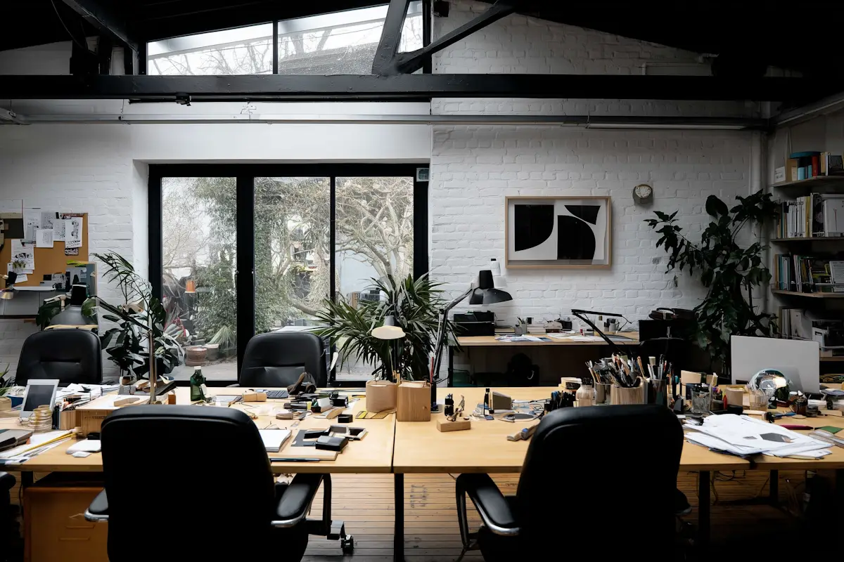 Modern office with wooden desks, black chairs, plants, large window, and office supplies.