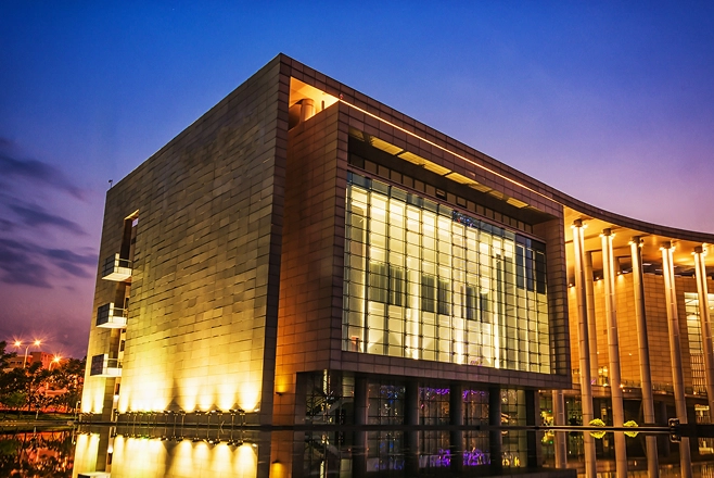 Modern building with large glass windows and columns illuminated at dusk reflecting on water.