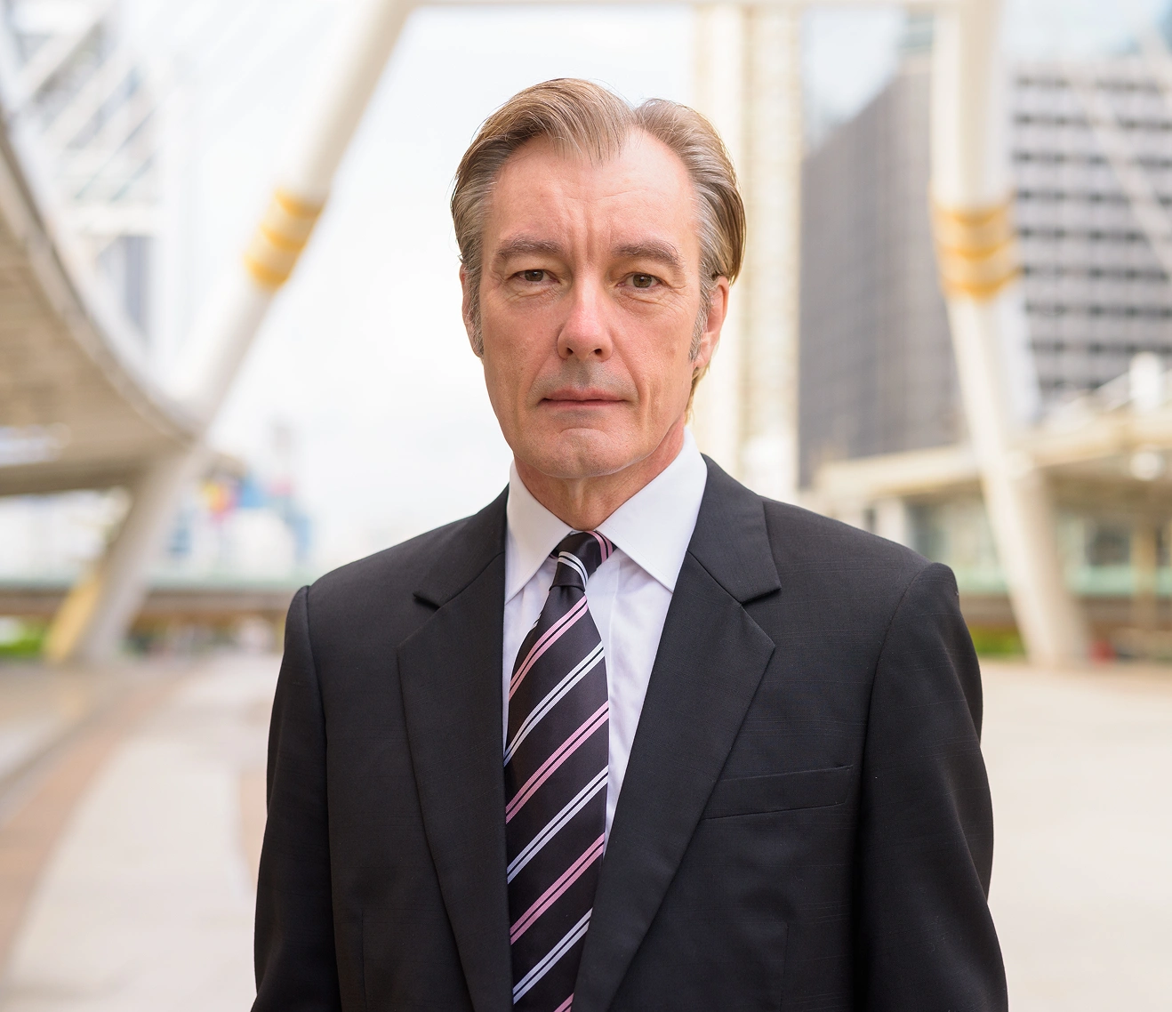 Middle-aged man in a dark suit and striped tie standing outdoors in an urban setting.