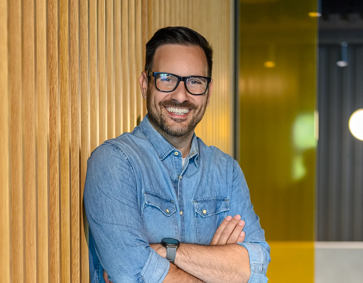 Smiling man with glasses and beard wearing a denim shirt, standing with arms crossed against a wooden wall.
