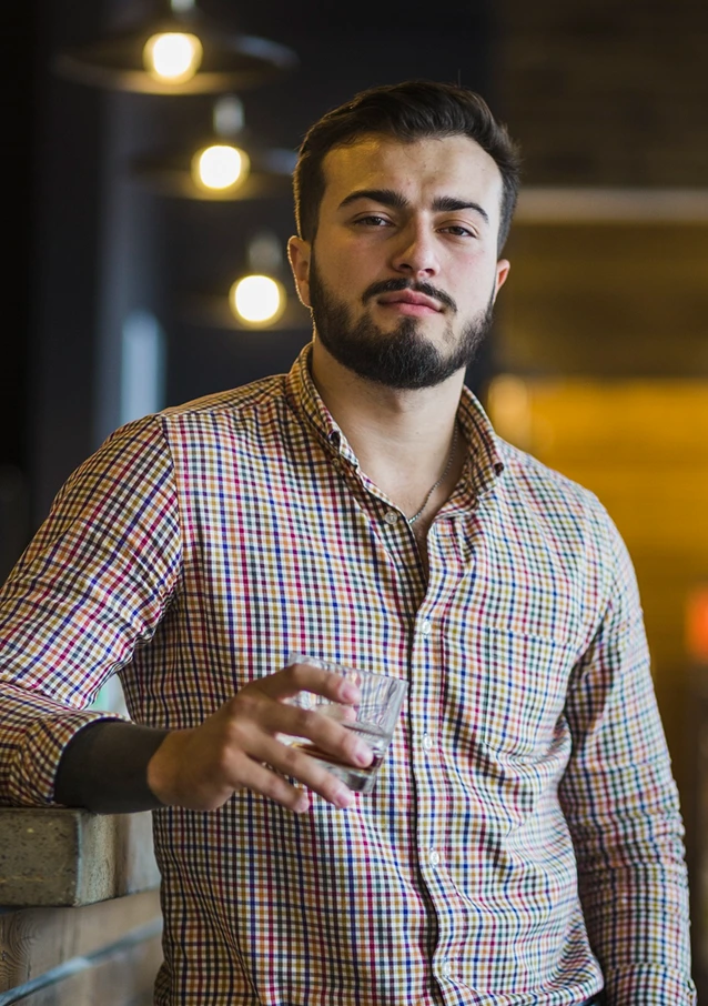 Bearded man in checkered shirt holding a glass, standing indoors with hanging lights in the background.