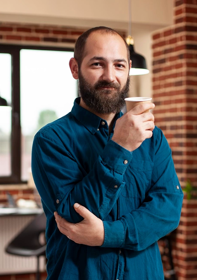 Bearded man in blue shirt holding a paper coffee cup in a cozy room with brick walls.