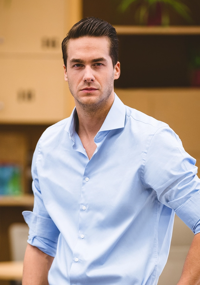 Confident young man with slicked-back hair wearing a light blue shirt with rolled-up sleeves.