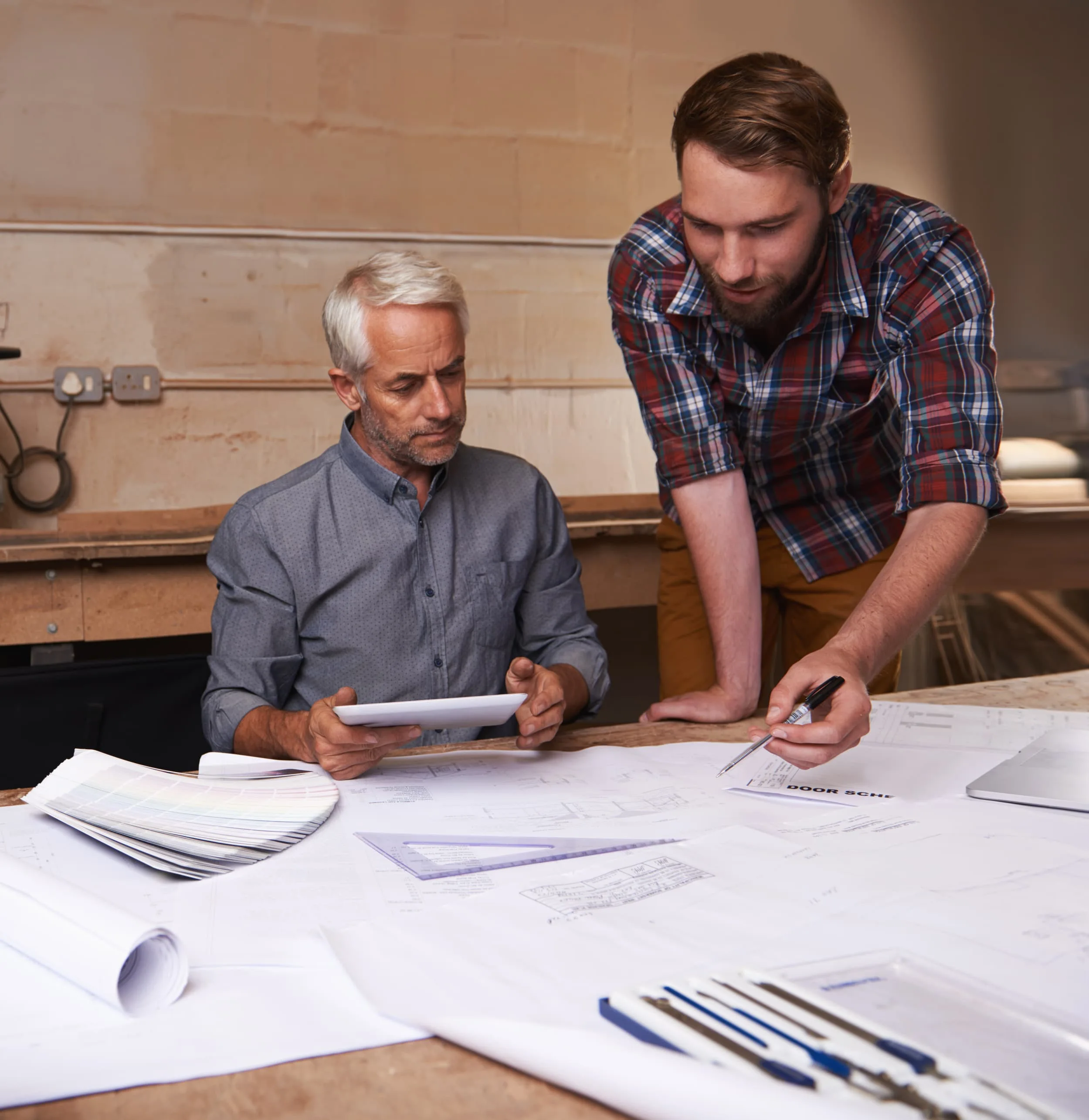 Two men studying architectural blueprints at a workspace with design tools and color swatches.