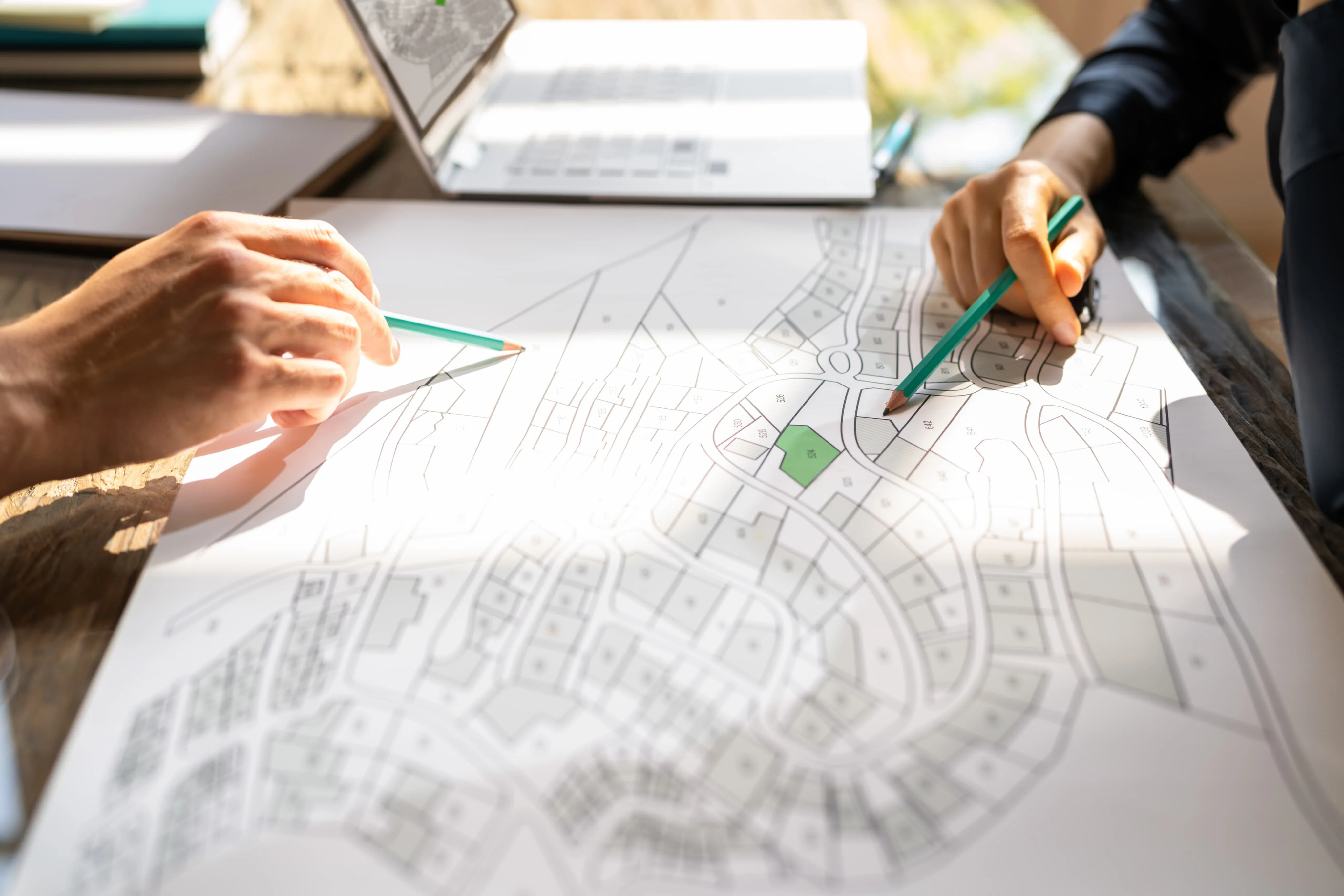 Two people pointing with green pencils at a residential map layout with marked lots on a table near a laptop.