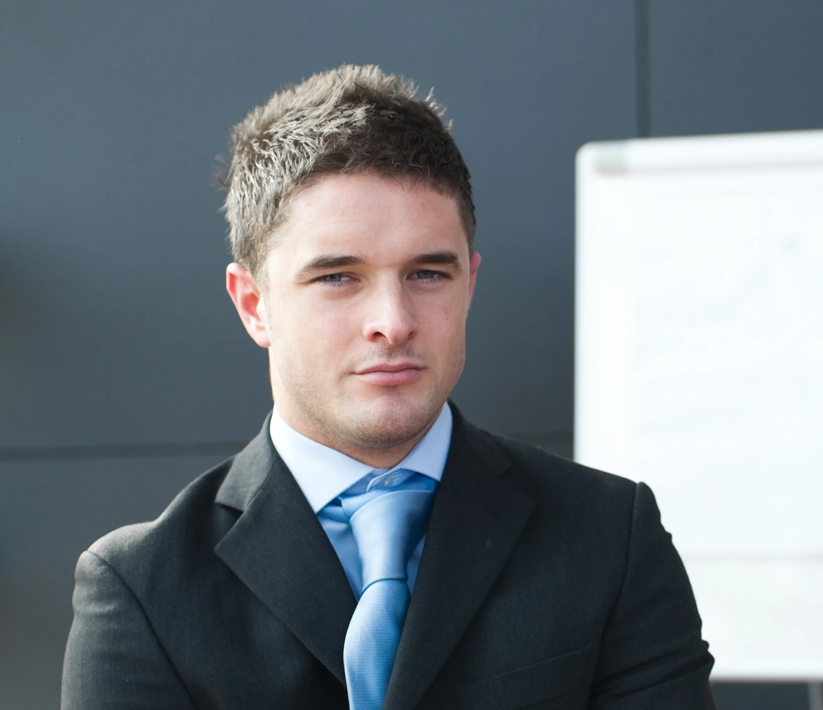 Confident young man in black suit and blue tie standing in front of a whiteboard.