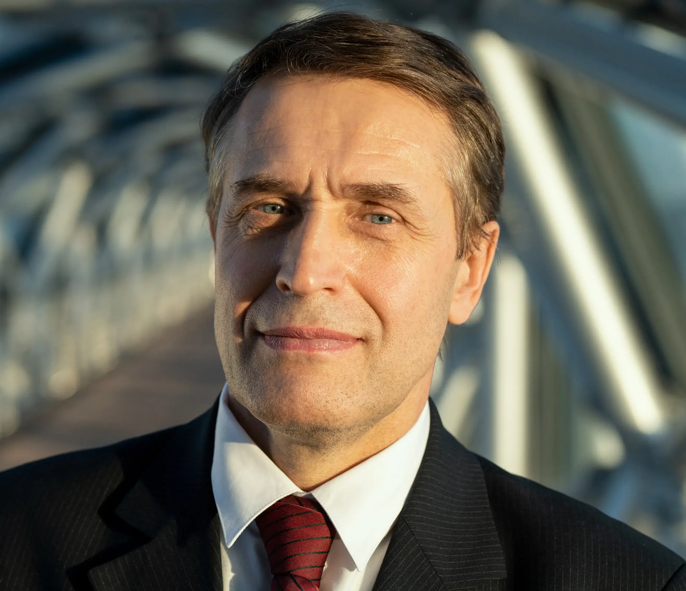 Middle-aged man in a suit and red tie standing outside in bright sunlight.