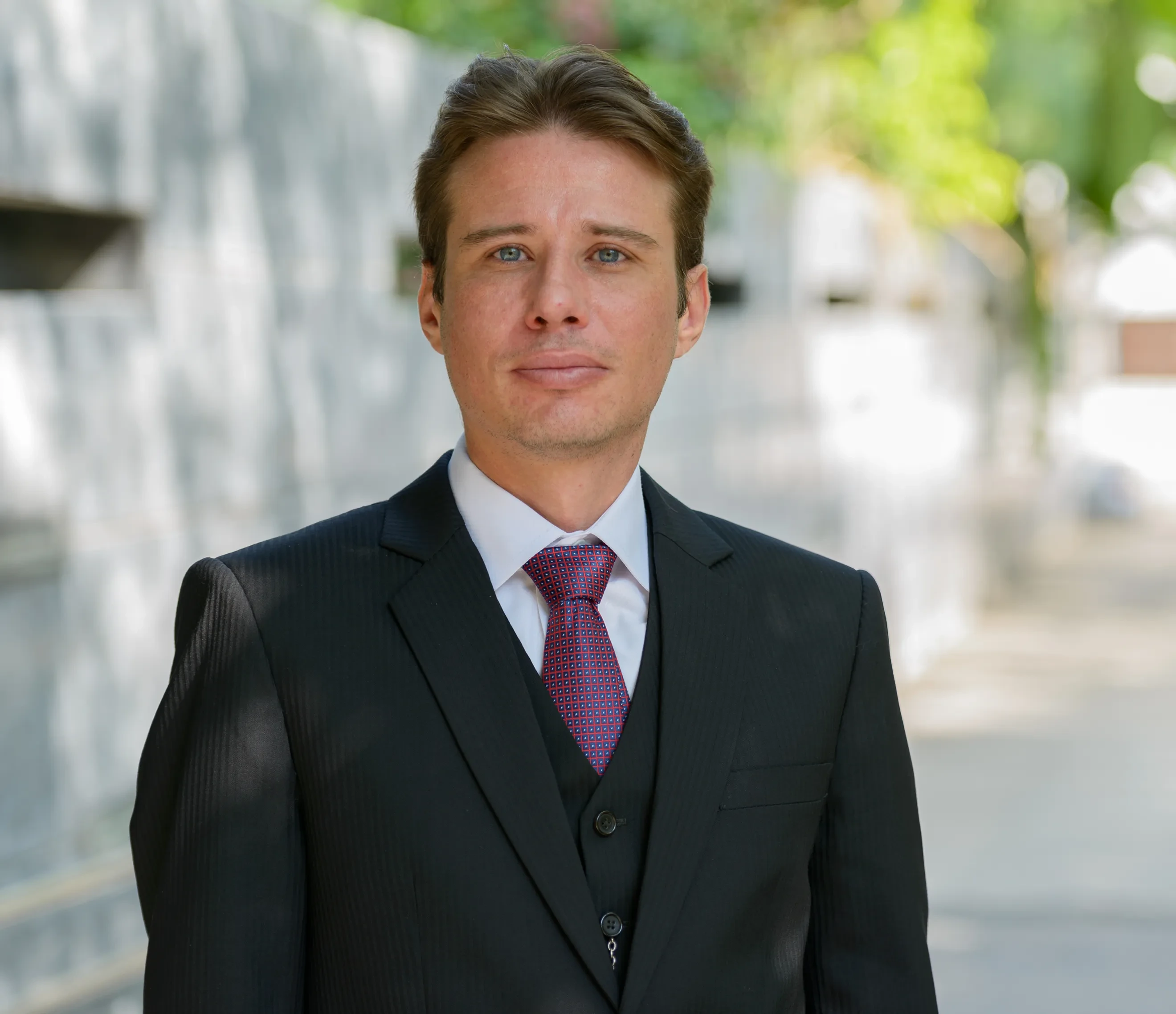 Man in black suit, white shirt, and red tie standing outdoors with blurred background.