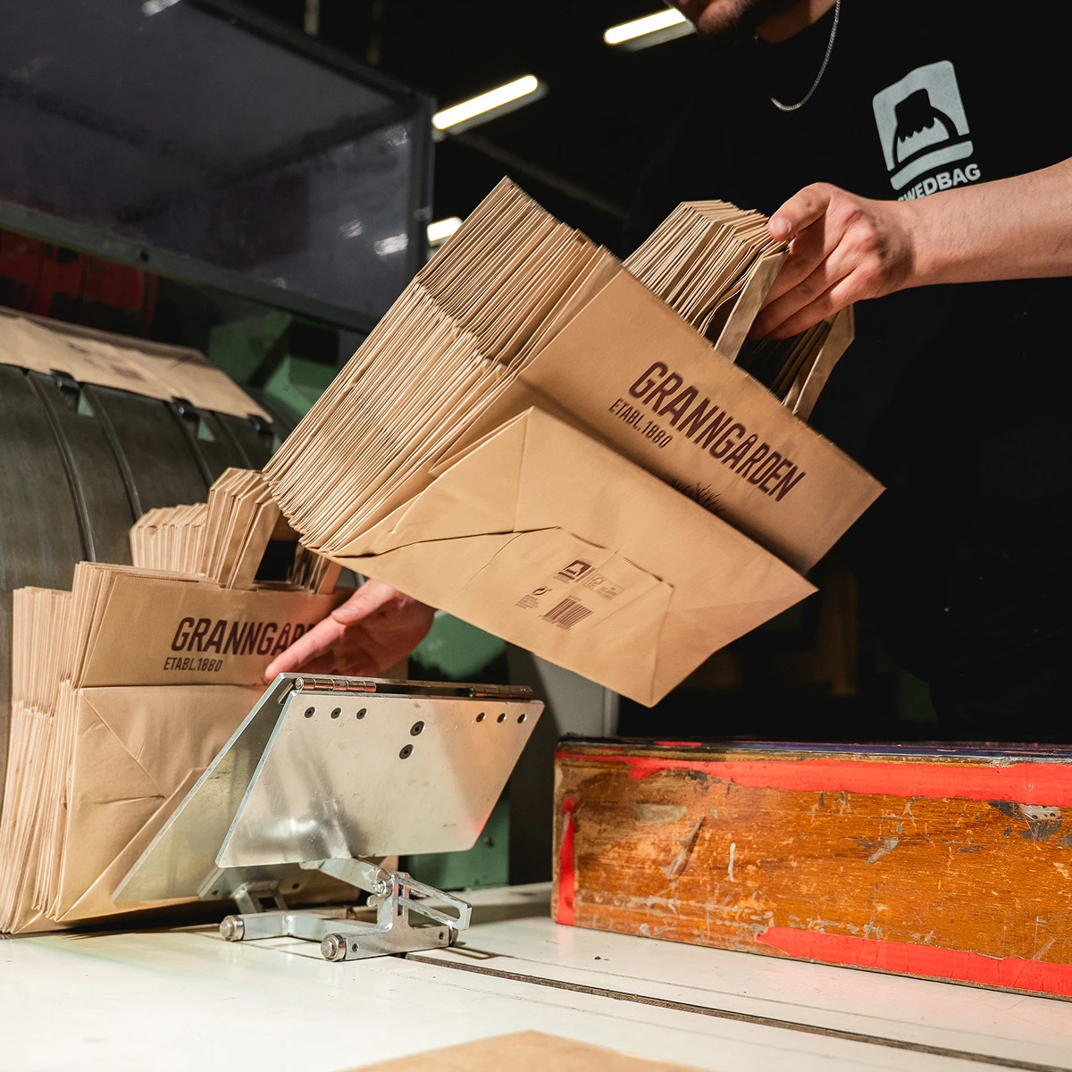 Person i swedbags fabrik som sorterar papperspåsar med texten Granngården i en industriell miljö.