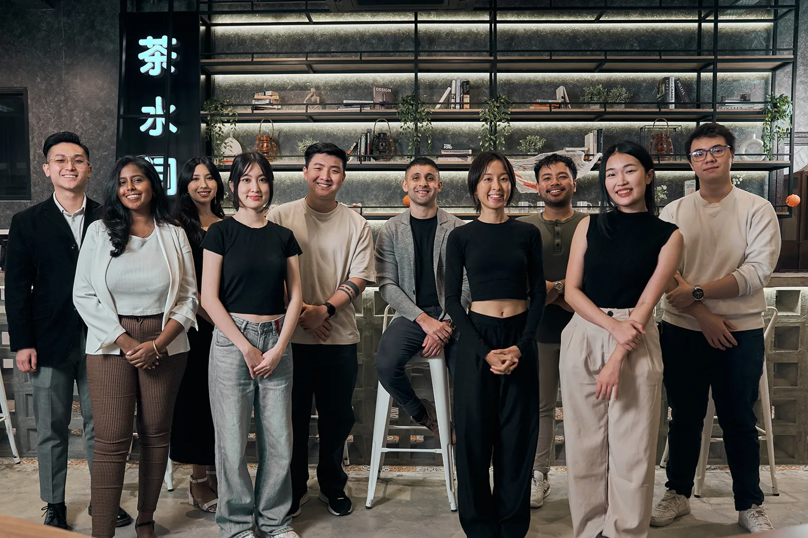 Smiling diverse group of ten young adults posing inside a modern cafe with shelves and plants behind.