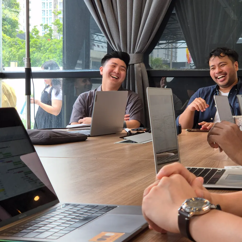 People laughing and chatting around a wooden table with laptops in a bright office setting.