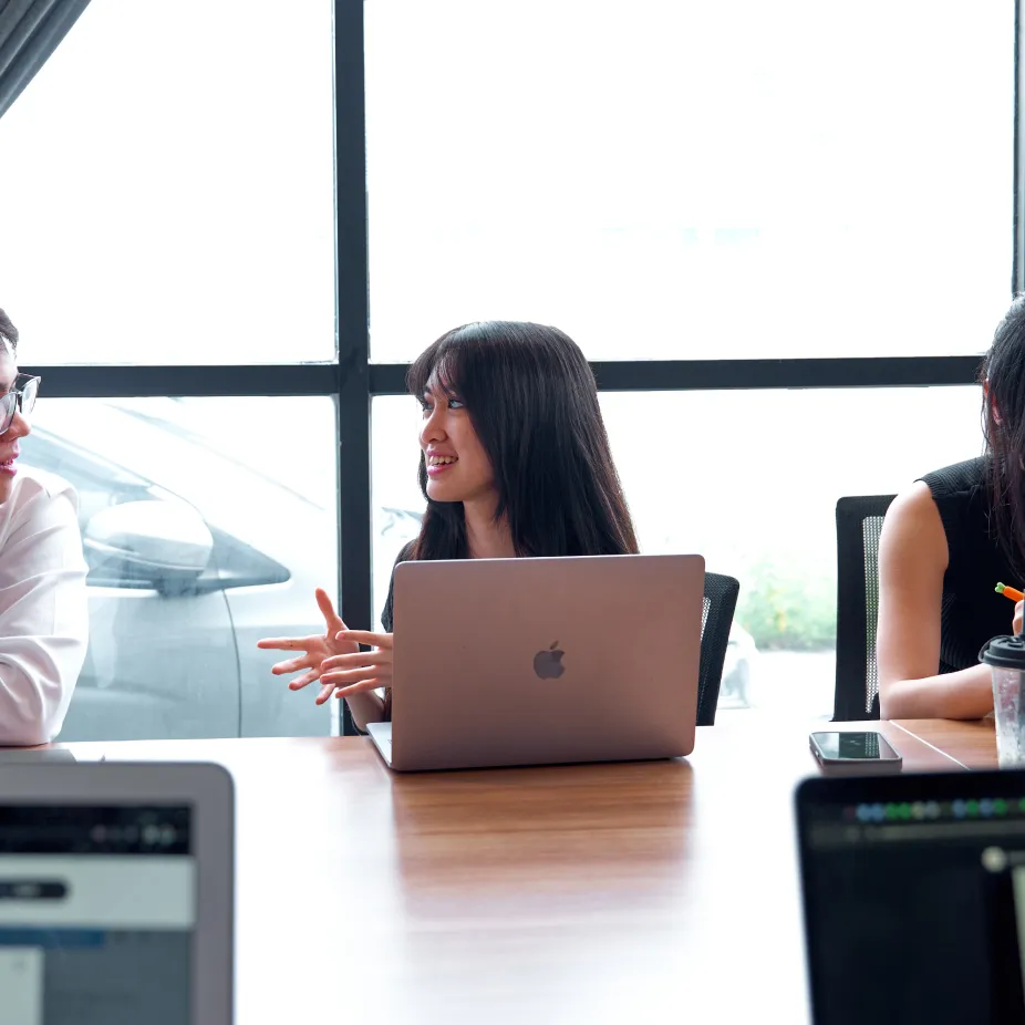 Young woman with long black hair talking and gesturing across a table with a laptop in front of her.