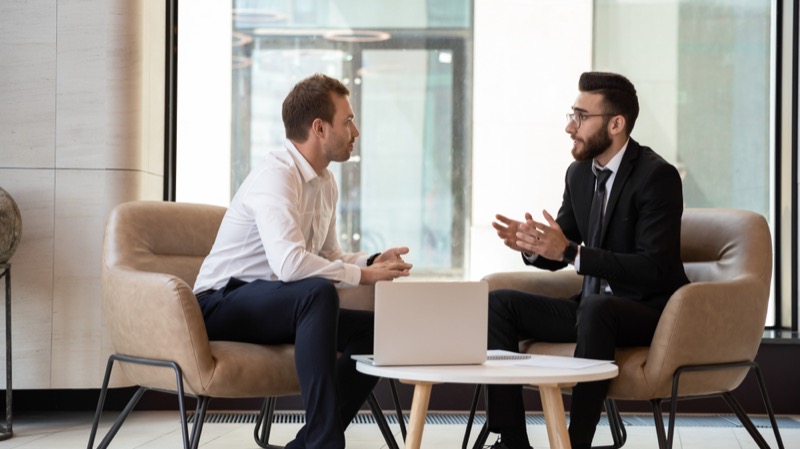 Middle eastern and caucasian ethnicity businessmen seated on armchair in modern office talking IT Consulting