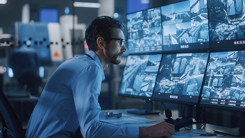 Industry 4.0 Modern Factory: Security Operator Controls Proper Functioning of Workshop Production Line, Uses Computer with Screens Showing Surveillance Camera Feed. High-Tech Security