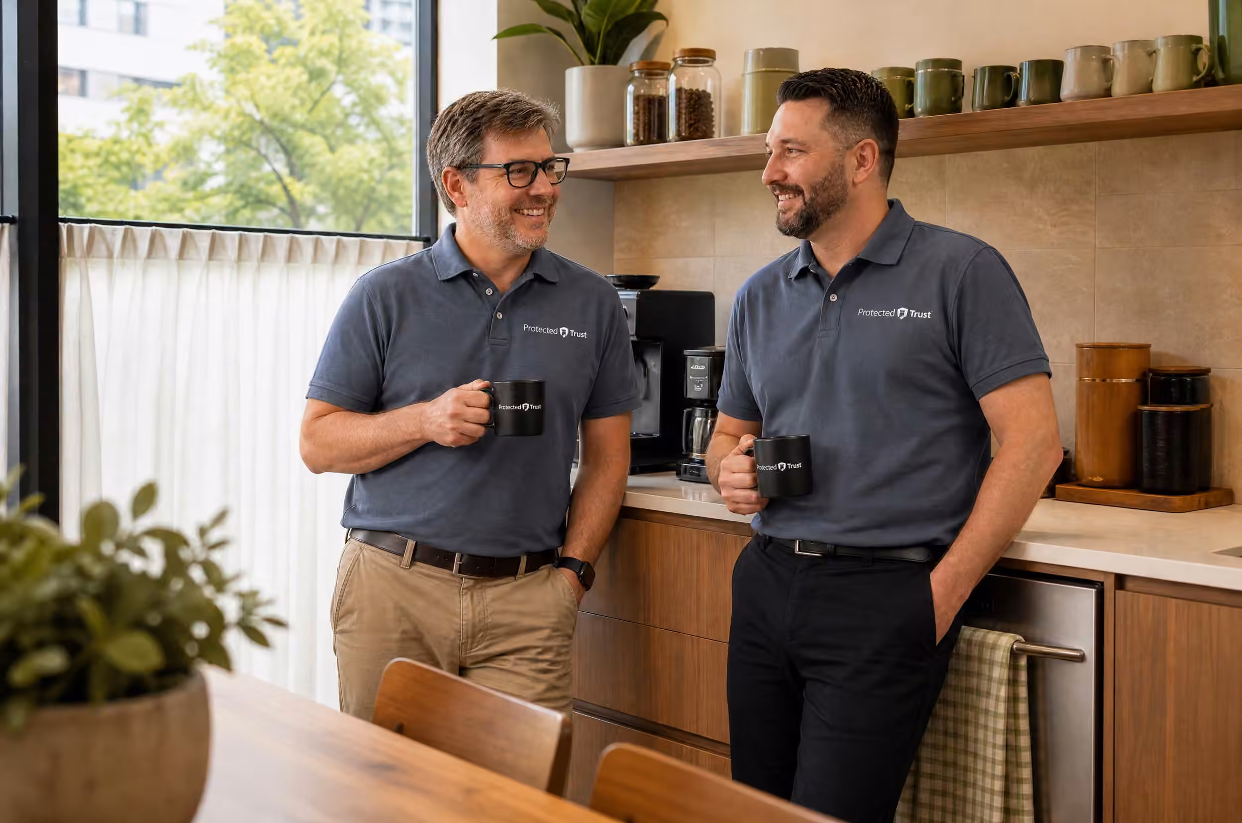 Two Protected Trust IT professionals chatting over coffee mugs in a modern office break room