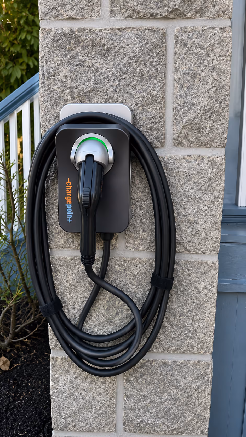 Electric vehicle charging station with coiled black cable mounted on a stone wall outside a house.